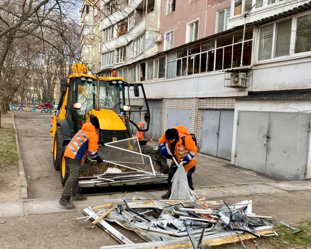 Municipal workers in high-vis jackets clear broken glass and twisted window frames outside a damaged apartment block