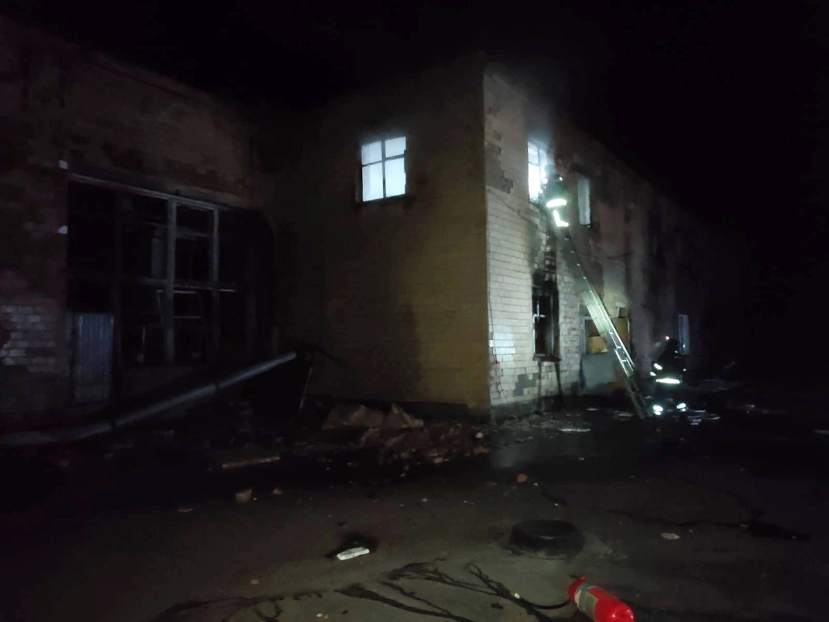 Firefighters climb a ladder to a lit upper-floor window of a damaged brick building at night